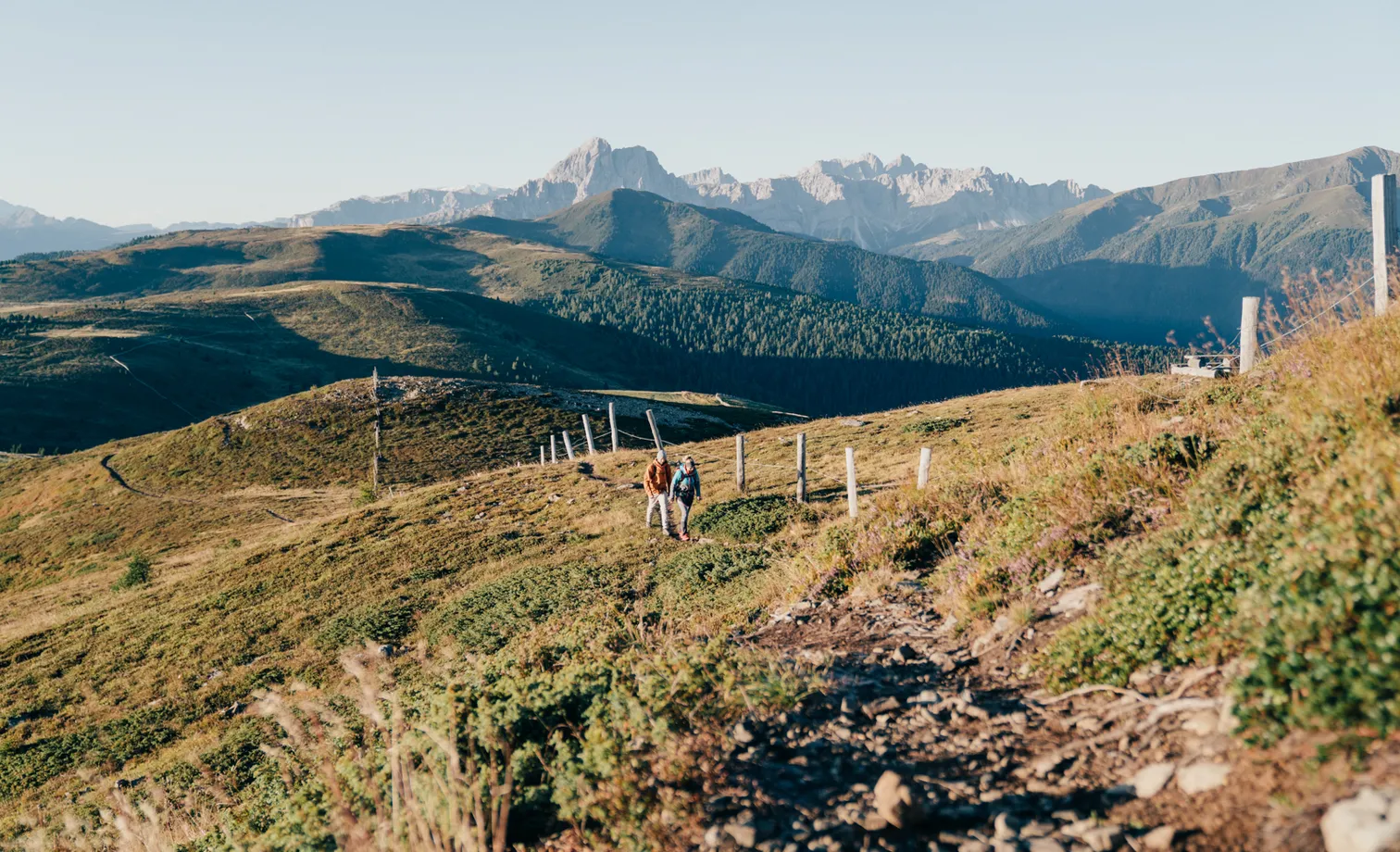 Two hikers on high trail above alpine meadows with panoramic view of the Geisler Dolomites in autumn near Luson