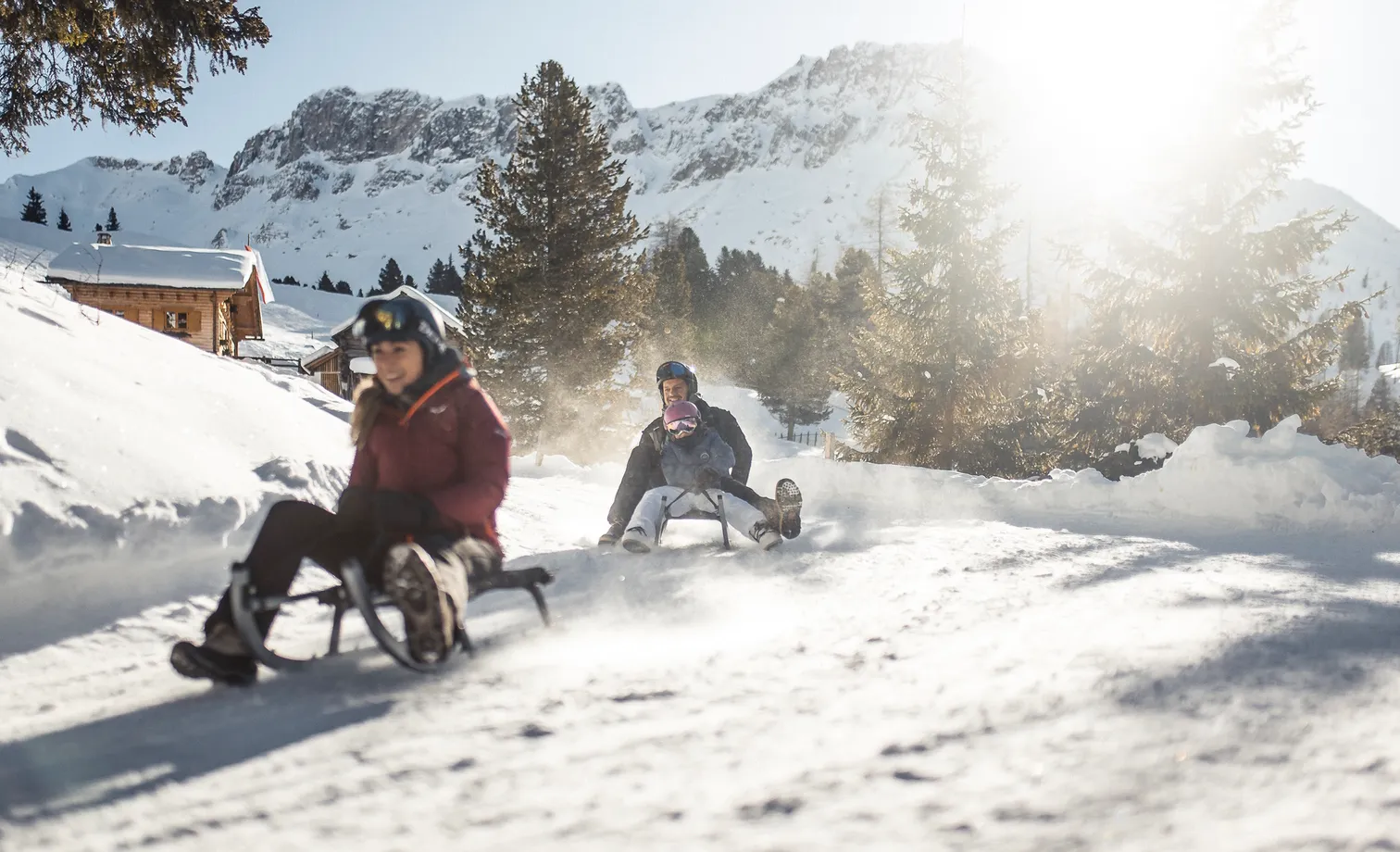 Family toboggans on wooden sled down sunny natural toboggan run, passing by alpine hut with Geisler group in backlight
