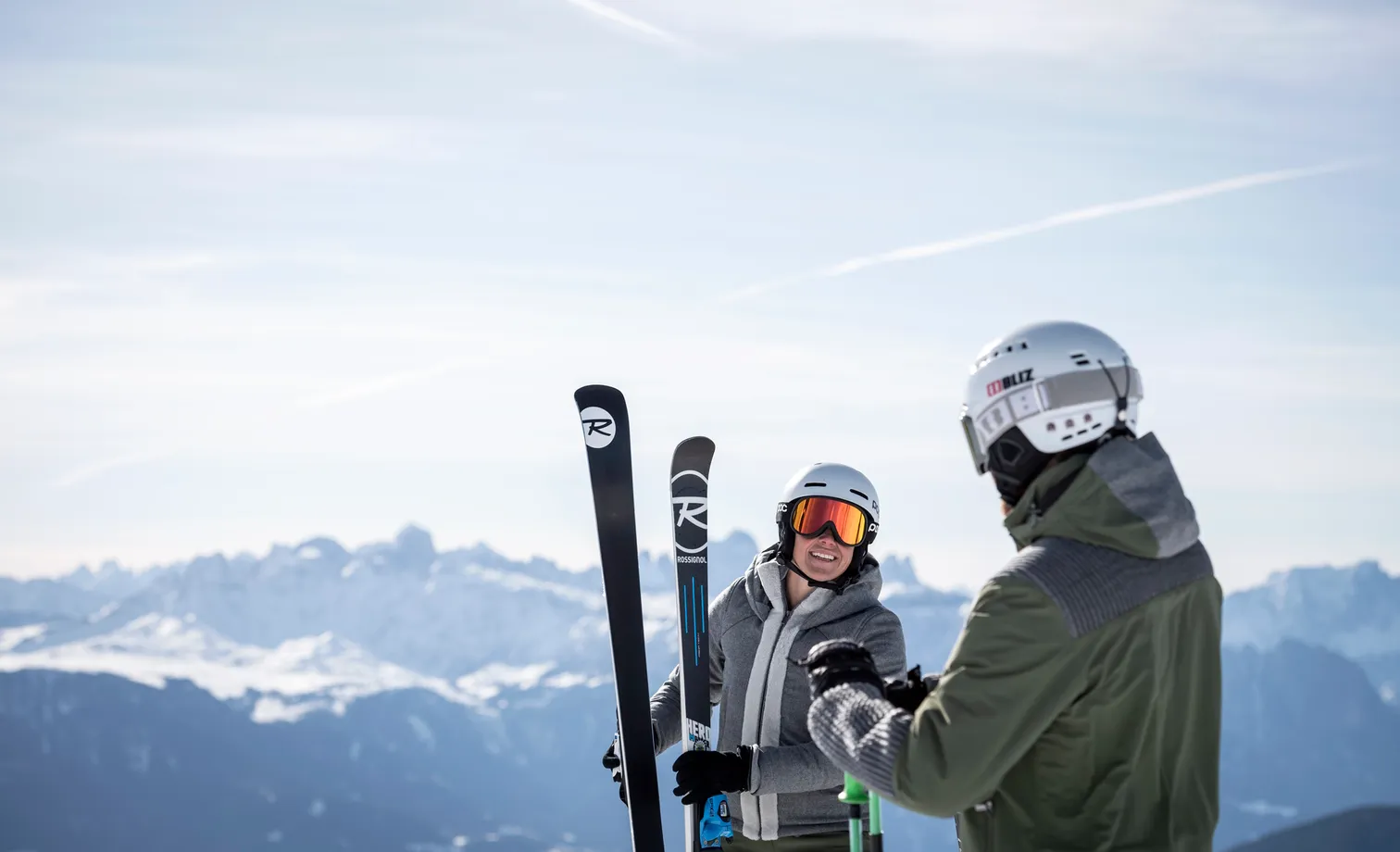 Smiling female skier with Rossignol skis and companion with Bliz helmet in front of snow-covered Dolomite panorama under clear sky in Villnöß