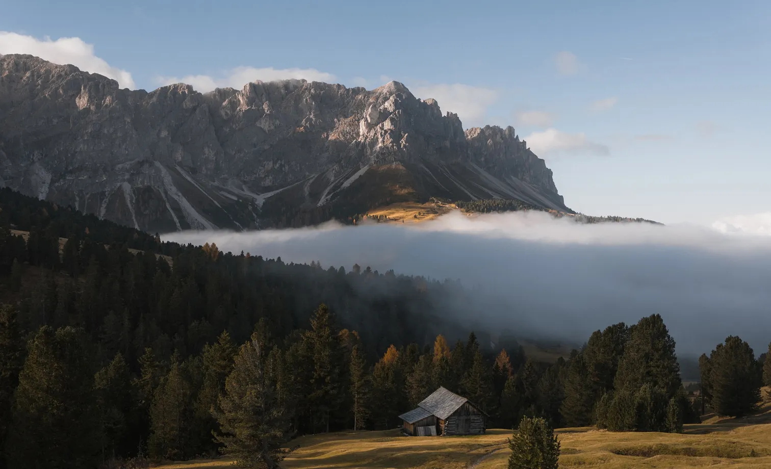 Autumn morning with fog band in front of Geisler group – wooden hut on alpine meadow surrounded by larches in Villnöß