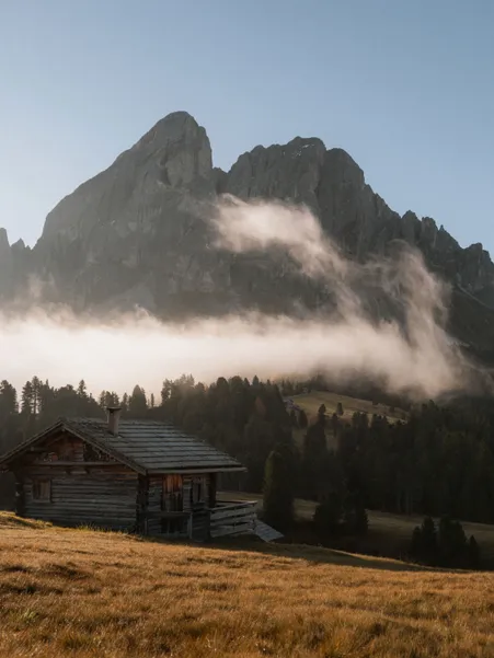 Wooden hut on alpine meadow in front of Peitlerkofel with dissolving morning fog in autumn