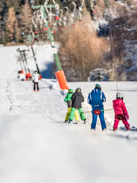 Children in colorful ski suits ride up on ski lift – ski school in sunshine with snow-covered forest in background