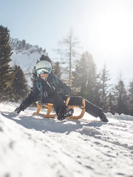 Boy brakes laughing with wooden sled in curve of toboggan run, Geisler peaks and backlight in background near Villnöß
