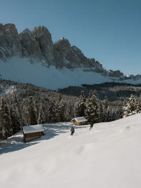 Ski tourer at wooden huts on snow-covered alpine meadow with view of Geisler group under clear winter sky in Villnöß