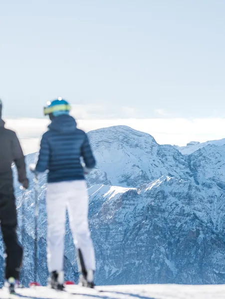 Two skiers from behind look towards the snow-covered peaks of the Geisler Dolomites – cloud band over the valley in clear winter light