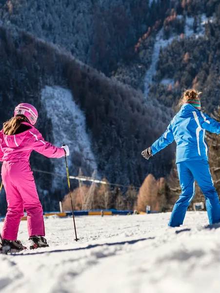 Ski instructor in blue suit shows girl in pink ski suit an exercise on flat slope in afternoon sun in Villnöß