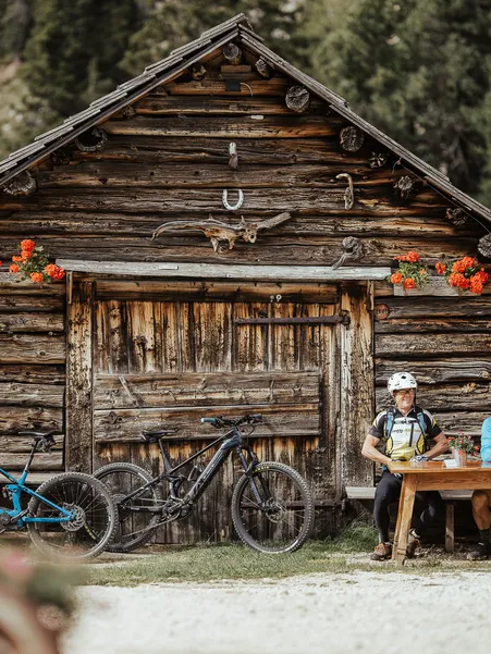 Mountain biker taking a break at rustic wooden hut with deer antlers and geraniums in Villnöß coniferous forest