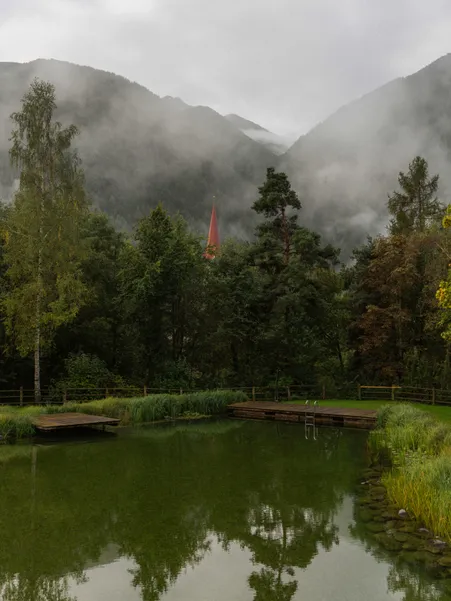 Natural bathing lake with wooden walkways and view of Luson bell tower in morning fog in front of South Tyrolean mountains
