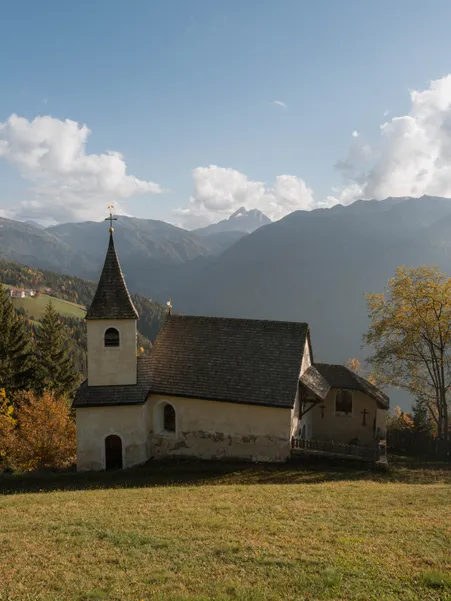 Historic chapel with shingle roof and bell tower on autumn meadow with view of mountain ranges near Luson in South Tyrol