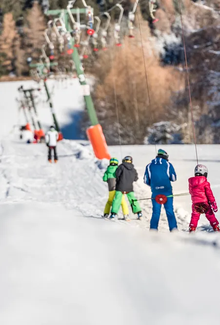 Kinder in bunten Skianzügen fahren am Schlepplift bergauf – Skischule bei Sonnenschein mit verschneitem Wald im Hintergrund