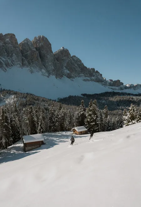 Skitourengeher bei Holzhütten auf verschneiter Almwiese mit Blick auf die Geislergruppe bei klarem Winterhimmel in Villnöß
