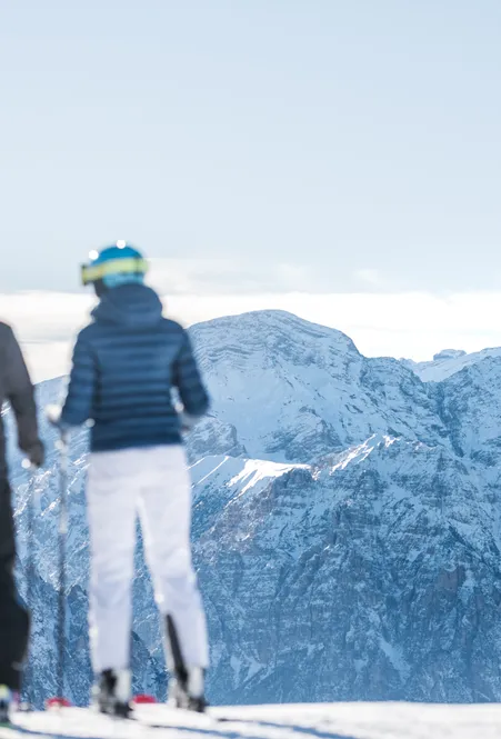 Zwei Skifahrer von hinten blicken auf die schneebedeckten Gipfel der Geisler-Dolomiten – Wolkenband über dem Tal bei klarem Winterlicht