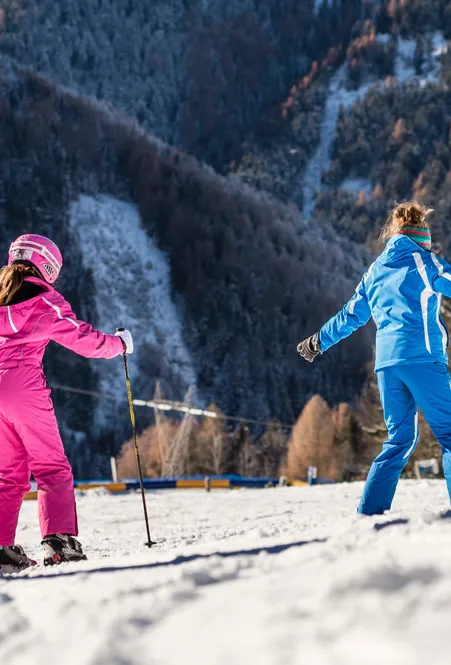 Skilehrerin in blauem Anzug zeigt Mädchen im pinken Skianzug eine Übung auf flacher Piste bei Nachmittagssonne in Villnöß