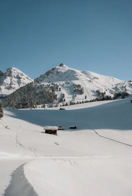 Tief verschneite Almlandschaft mit Holzhütten und Skitourenspuren vor sonnenbeschienenen Lüsner Bergkuppen bei blauem Himmel